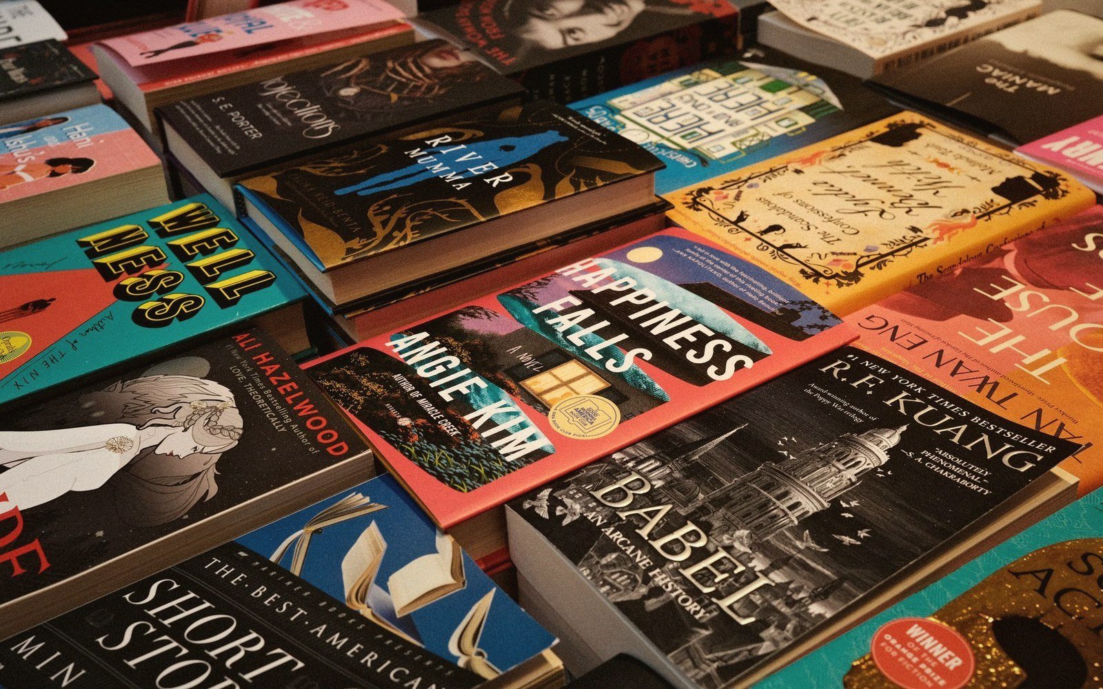 Stacks of colorful book covers overlapping on a table, including "Happiness Falls" and "Babel"
