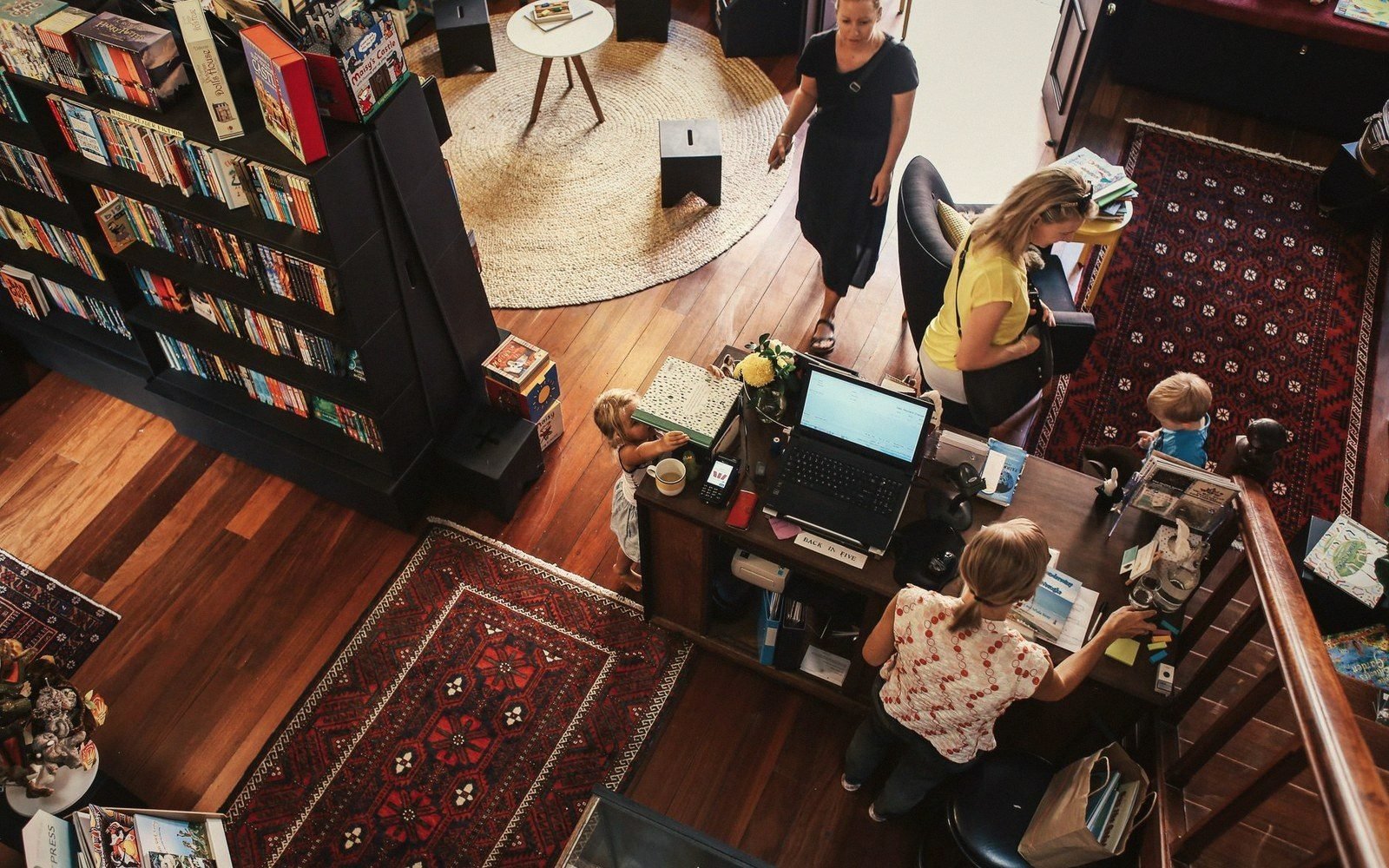 Overhead view of shoppers and staff at a bookshop counter with patterned rugs on wooden floors
