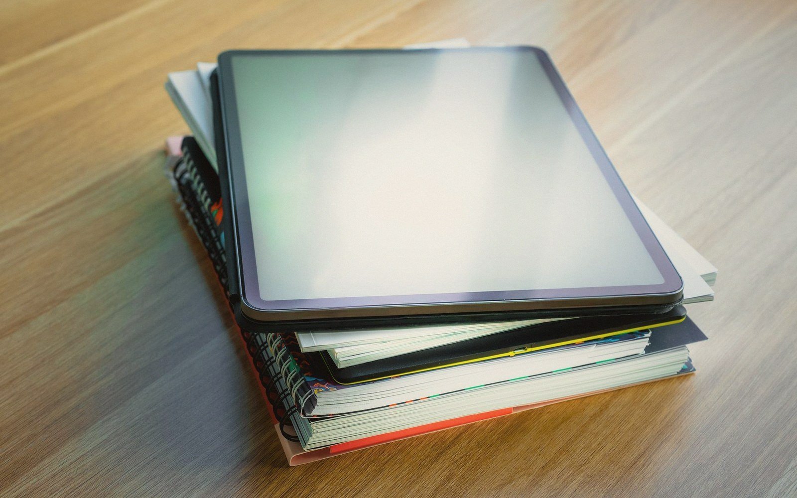 A tablet with a blank screen resting on a stack of notebooks on a wooden table