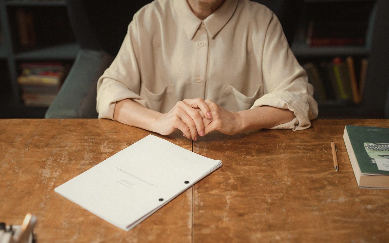 Person sits with clasped hands behind a bound manuscript on a wooden desk